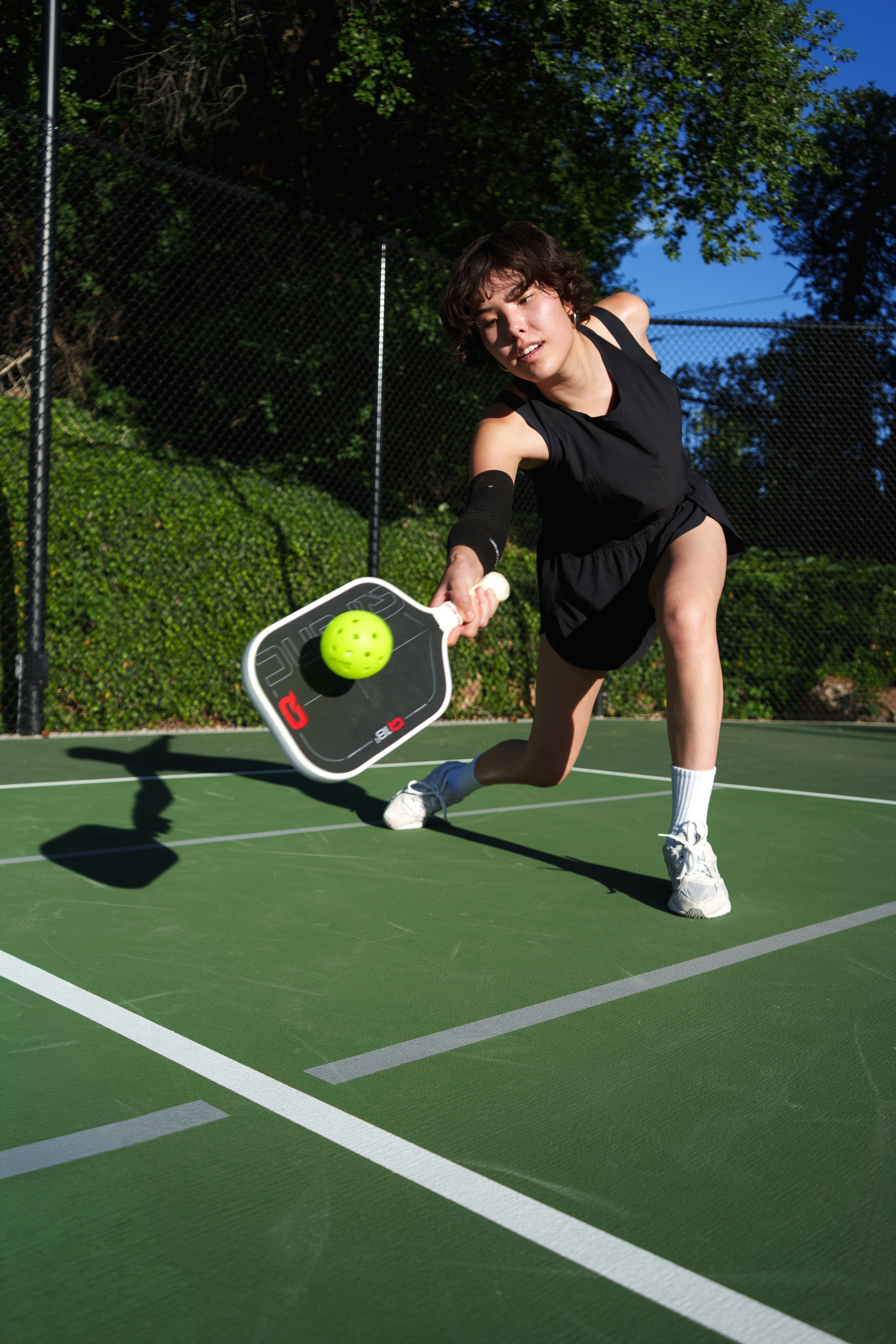 A woman in a black athletic outfit plays pickleball, swinging a paddle with the Q-ball in mid-air.